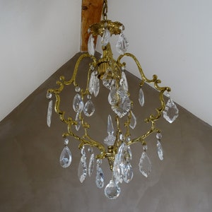 Puede incluir: Una l&aacute;mpara de ara&ntilde;a de lat&oacute;n vintage con m&uacute;ltiples colgantes de cristal transparente. La l&aacute;mpara de ara&ntilde;a tiene un dise&ntilde;o ornamental con brazos curvos y una columna central. Los cristales son de varias formas, incluyendo l&aacute;grimas y piezas facetadas, reflejando la luz.