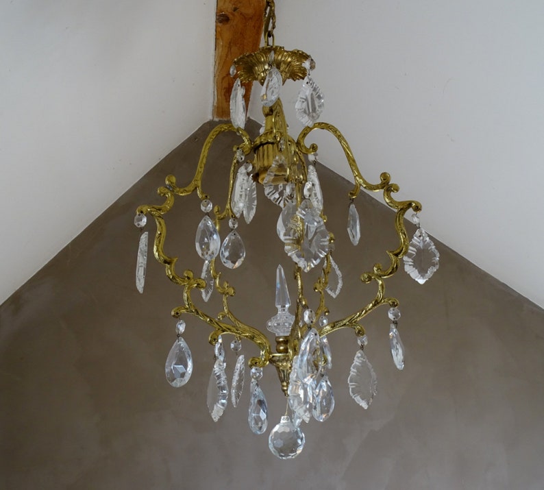 Puede incluir: Una l&aacute;mpara de ara&ntilde;a de lat&oacute;n vintage con m&uacute;ltiples colgantes de cristal transparente. La l&aacute;mpara de ara&ntilde;a tiene un dise&ntilde;o ornamental con brazos curvos y una columna central. Los cristales son de varias formas, incluyendo l&aacute;grimas y piezas facetadas, reflejando la luz.