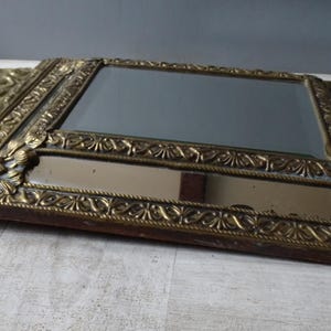 Puede incluir: Un espejo rectangular antiguo con un marco dorado ornamentado. El marco presenta detalles florales y de volutas. El espejo descansa sobre una superficie de color claro, mostrando su est&eacute;tica vintage.