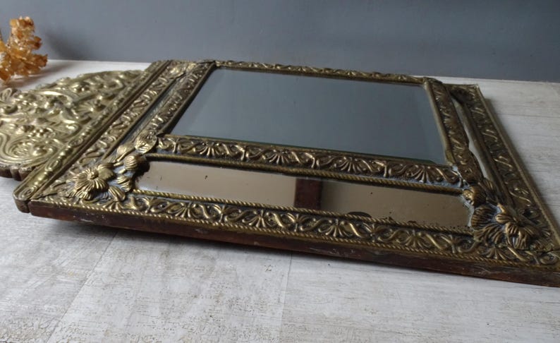 Puede incluir: Un espejo rectangular antiguo con un marco dorado ornamentado. El marco presenta detalles florales y de volutas. El espejo descansa sobre una superficie de color claro, mostrando su est&eacute;tica vintage.