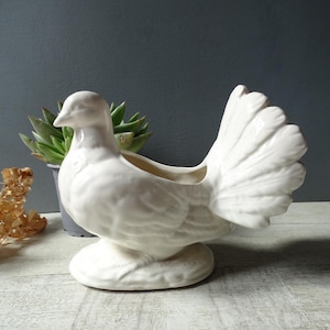 Puede incluir: Una maceta de cerámica blanca con forma de paloma, con una cola detallada y una base redonda. La paloma está posada sobre una base, y la maceta tiene una abertura en la parte posterior para plantas. Es un objeto decorativo.
