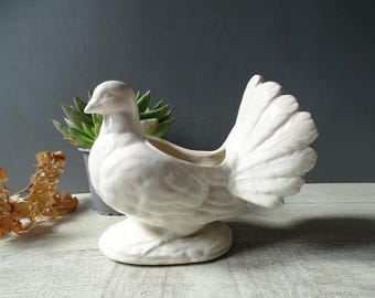 May include: A white ceramic dove planter with a detailed tail and a round base. The dove is perched on a base, and the planter has an opening on its back for plants. The dove is a decorative item.