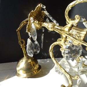Puede incluir: Primer plano de una l&aacute;mpara de ara&ntilde;a de lat&oacute;n vintage con detalles de cristal transparente. La l&aacute;mpara de ara&ntilde;a presenta brazos curvos y ornamentados y una base en forma de campana. Los cristales son de varias formas, incluyendo l&aacute;grimas y piezas facetadas.