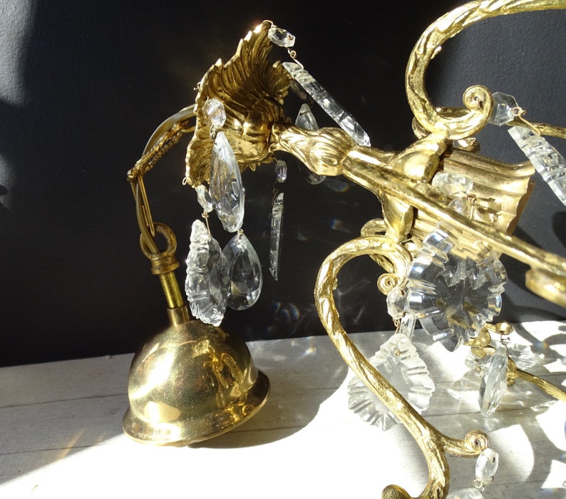 Puede incluir: Primer plano de una l&aacute;mpara de ara&ntilde;a de lat&oacute;n vintage con detalles de cristal transparente. La l&aacute;mpara de ara&ntilde;a presenta brazos curvos y ornamentados y una base en forma de campana. Los cristales son de varias formas, incluyendo l&aacute;grimas y piezas facetadas.