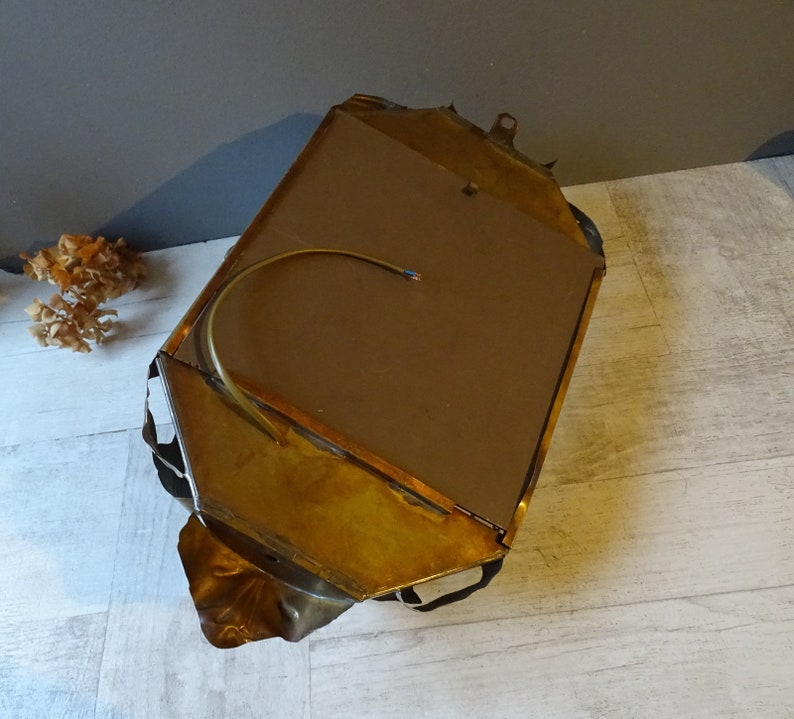 May include: A gold-toned metal wall mirror with a unique, abstract design. The mirror has a brown backing and a wire hanging loop.
