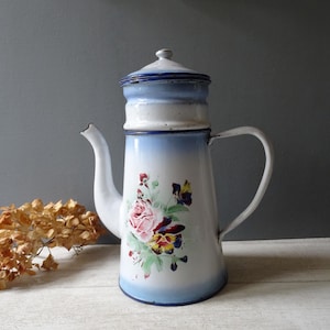 Könnte beinhalten: Vintage Emaille-Kaffeekanne mit weißem Korpus, hellblauem Deckel und weißem Griff. Die Kanne hat ein Blumendesign mit rosa Rosen und violetten Stiefmütterchen. Die Kanne ist ca. 25 cm hoch.
