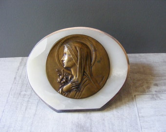 May include: A white oval-shaped frame with a bronze relief of a woman's profile. The woman is wearing a veil and holding a flower.