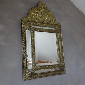 Puede incluir: Espejo dorado ornamentado, de forma rectangular, con detalles decorativos. El marco presenta dise&ntilde;os florales y de volutas. La parte superior del espejo tiene un escudo elaborado. Montado sobre una pared gris.