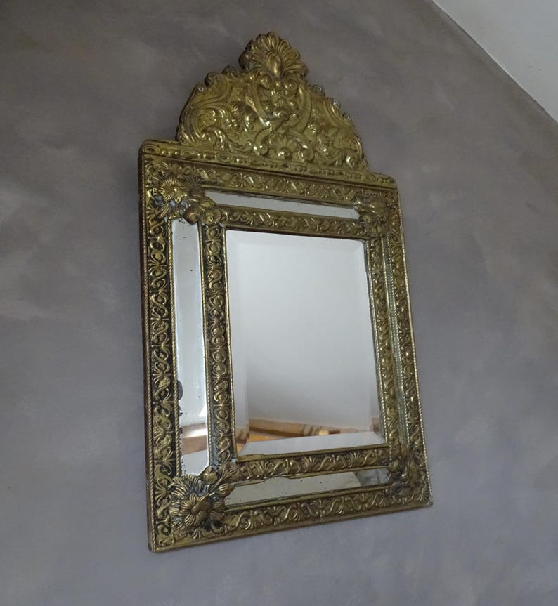 Puede incluir: Espejo dorado ornamentado, de forma rectangular, con detalles decorativos. El marco presenta dise&ntilde;os florales y de volutas. La parte superior del espejo tiene un escudo elaborado. Montado sobre una pared gris.