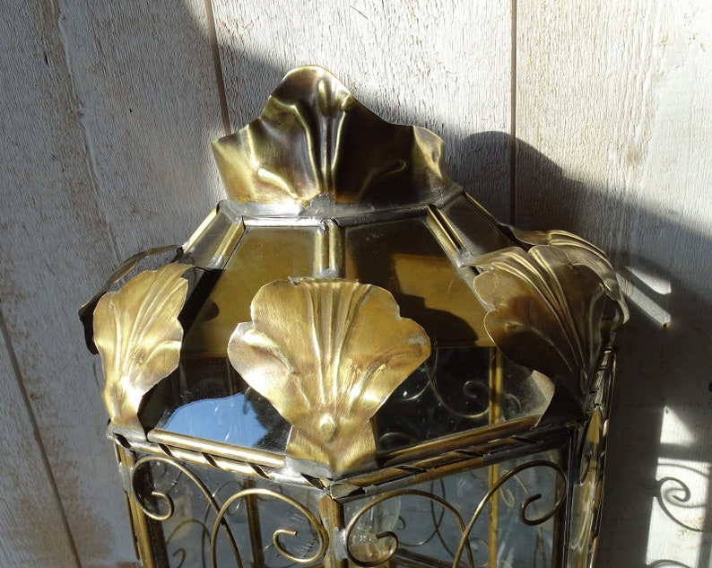 May include: A gold metal lantern with a glass top and ornate metal detailing. The lantern has a decorative design with a floral motif.
