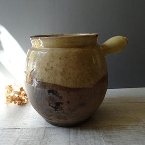 Puede incluir: Una olla grande, rústica, de cerámica esmaltada con una sola asa. La olla es marrón y beige con un esmalte moteado. La olla está sobre una superficie de madera clara.