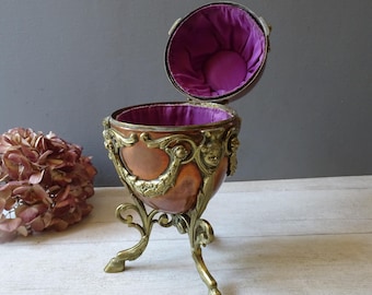 May include: An antique egg-shaped trinket box with a copper body and ornate brass detailing. The box is open, revealing a purple velvet interior. The box sits on three ornate brass legs.