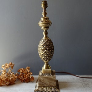 Puede incluir: Una lámpara de mesa dorada con una base en forma de piña y una parte superior decorativa. La lámpara tiene una base de mármol y un cable marrón.