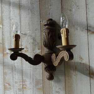 Pode incluir: Arandela de parede castanho escuro com duas luzes e detalhes esculpidos ornamentados. A arandela apresenta duas lâmpadas em forma de vela com gotejamento de cera falso. A luminária é montada em uma parede de madeira clara.