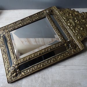 Puede incluir: Un espejo dorado antiguo con un marco ornamentado y detallado. El espejo rectangular tiene una superficie oscura y reflectante y est&aacute; en &aacute;ngulo, mostrando el intrincado dise&ntilde;o floral y de volutas del marco. El espejo es un art&iacute;culo decorativo.