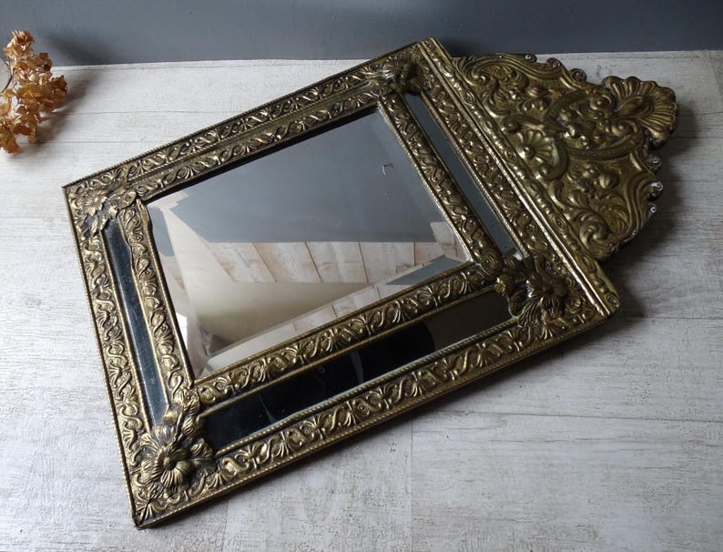 Puede incluir: Un espejo dorado antiguo con un marco ornamentado y detallado. El espejo rectangular tiene una superficie oscura y reflectante y est&aacute; en &aacute;ngulo, mostrando el intrincado dise&ntilde;o floral y de volutas del marco. El espejo es un art&iacute;culo decorativo.