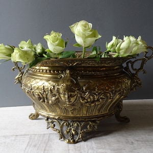 May include: A large, ornate brass planter with intricate details and scrollwork. The planter has two handles and sits on three feet. It is filled with white roses.