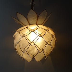 Op de afbeelding: Een decoratieve hanglamp in de vorm van een ananas. Het licht bestaat uit witte, bloembladachtige segmenten die een warme gloed uitstralen. De lamp hangt aan een ketting en biedt een stijlvolle verlichtingsoplossing.
