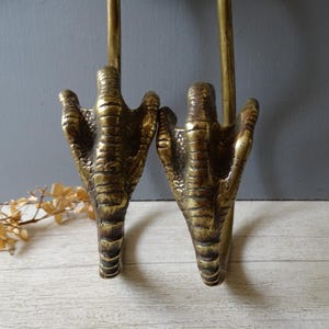 May include: A pair of brass wall hooks shaped like bird talons. Each hook features a textured, three-toed claw design with a metallic gold finish. The hooks are curved and extend outwards, showing detailed scales and joints on the surface of the metal.