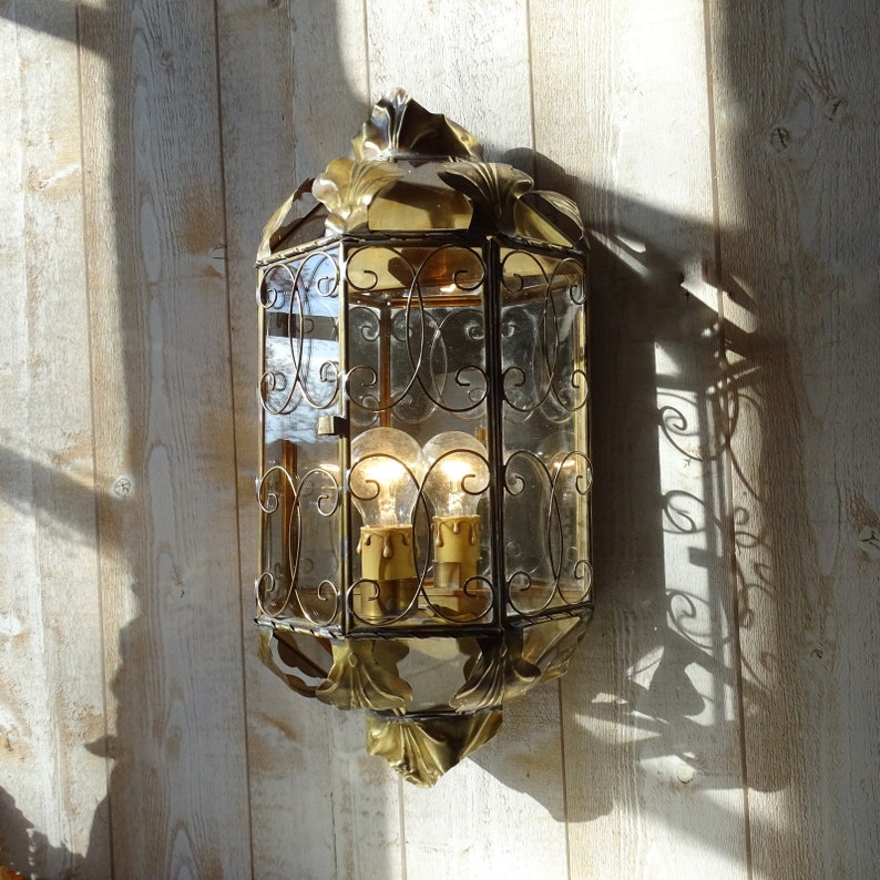 May include: A gold-toned metal lantern with a glass pane and intricate scrollwork. The lantern has two light bulbs inside.