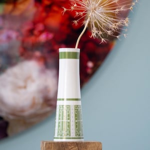 Può includere: Un vaso di ceramica bianco con motivi geometrici e strisce verdi. Il vaso contiene un fiore secco dall'aspetto delicato e arioso. Il vaso è posto su una superficie di legno contro uno sfondo sfocato rosso e blu.