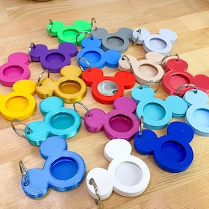 May include: A collection of colorful Mickey Mouse-shaped keychains in various colors, including green, purple, blue, pink, yellow, and red. Each keychain has a circular opening and a silver keyring. The keychains are arranged on a light brown wooden surface.