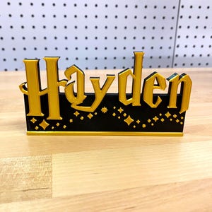 May include: A black and gold name sign with the name "Hayden" in a decorative font. The sign has a black background with gold stars and diamonds.
