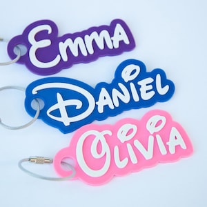May include: Three colorful personalized keychains with metal rings and cable chains. The keychains are purple, blue, and pink and have the names "Emma", "Daniel", and "Olivia" written in white.