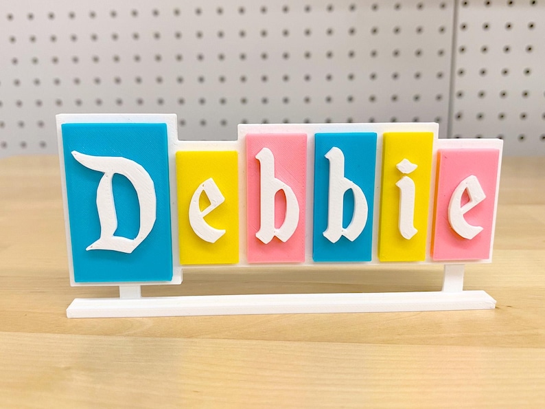 May include: A white nameplate with the name "Debbie" spelled out in colourful blocks. The blocks are blue, yellow, pink, blue, yellow, and pink. The nameplate is on a wooden surface.