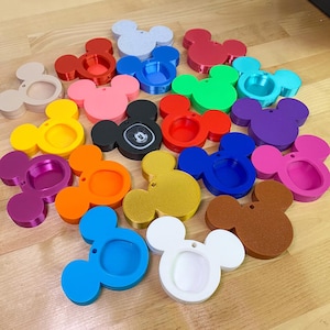 May include: A collection of colorful Mickey Mouse-shaped trinket holders in various colors, including red, blue, green, and yellow. Some have a textured appearance, while others are smooth. The holders are arranged on a light-colored wooden surface.