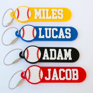 May include: Four personalized keychains with baseball-themed charms and names "MILES", "LUCAS", "ADAM", and "JACOB" printed on them. The keychains are in yellow, blue, black, and red.