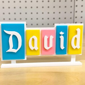 May include: A white plastic name sign with the name "David" spelled out in white letters on colourful blocks. The blocks are blue, yellow, pink, blue, and yellow.