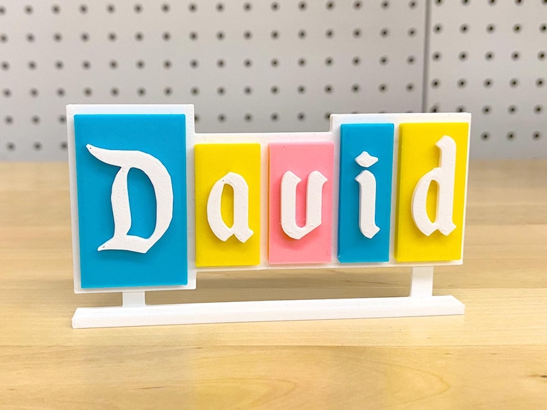 May include: A white plastic name sign with the name "David" spelled out in white letters on colourful blocks. The blocks are blue, yellow, pink, blue, and yellow.
