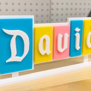 May include: A white plastic name sign with the name "David" spelled out in white letters on coloured blocks. The blocks are blue, yellow, pink, and blue. The sign is on a white stand.