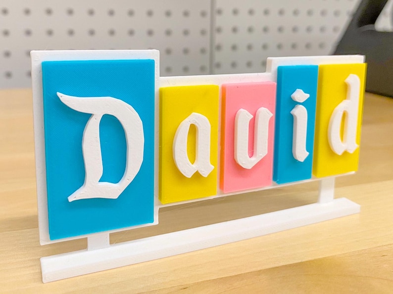 May include: A white plastic name sign with the name "David" spelled out in white letters on coloured blocks. The blocks are blue, yellow, pink, and blue. The sign is on a white stand.