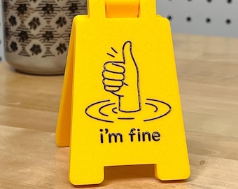 May include: A bright yellow sign with a black outline of a hand giving a thumbs up, emerging from water. The words "i'm fine" are printed in black below the graphic. The sign is a two-sided, folding design.