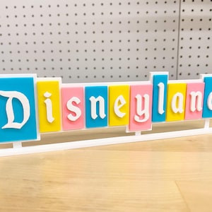 May include: A colourful sign spelling out "Disneyland" in white letters on a blue, yellow, pink and turquoise background. The sign is on a white stand.