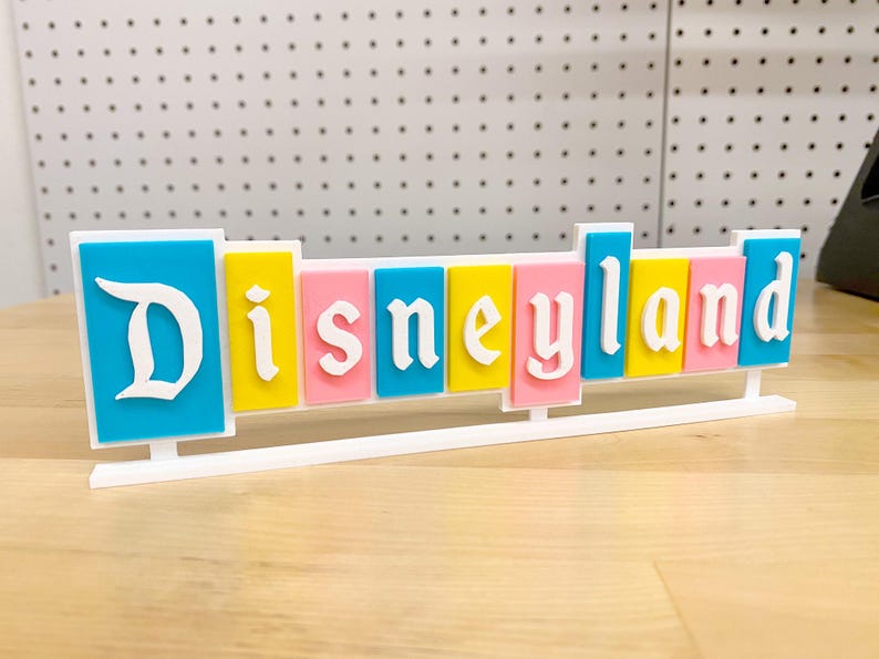 May include: A colourful sign spelling out "Disneyland" in white letters on a blue, yellow, pink and turquoise background. The sign is on a white stand.