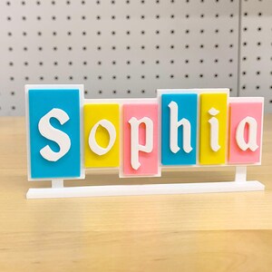 May include: A white stand with the name "Sophia" spelled out in colourful blocks. The blocks are blue, yellow, pink, and light blue.