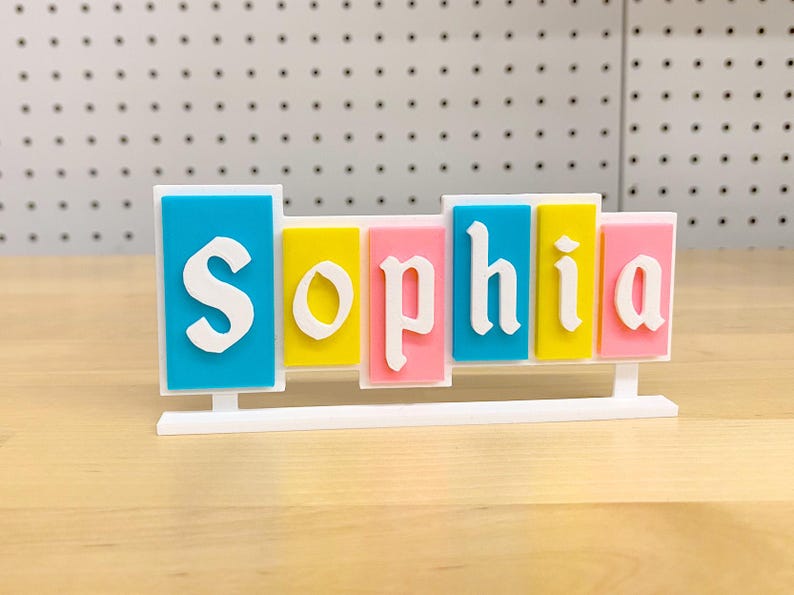 May include: A white stand with the name "Sophia" spelled out in colourful blocks. The blocks are blue, yellow, pink, and light blue.