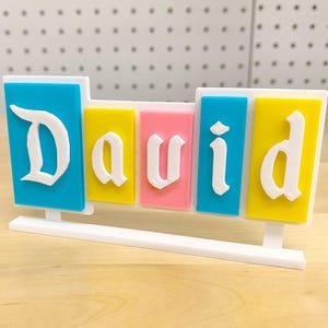 May include: A white nameplate with the name "David" spelled out in white letters on colourful blocks. The blocks are blue, yellow, pink, blue, and yellow.