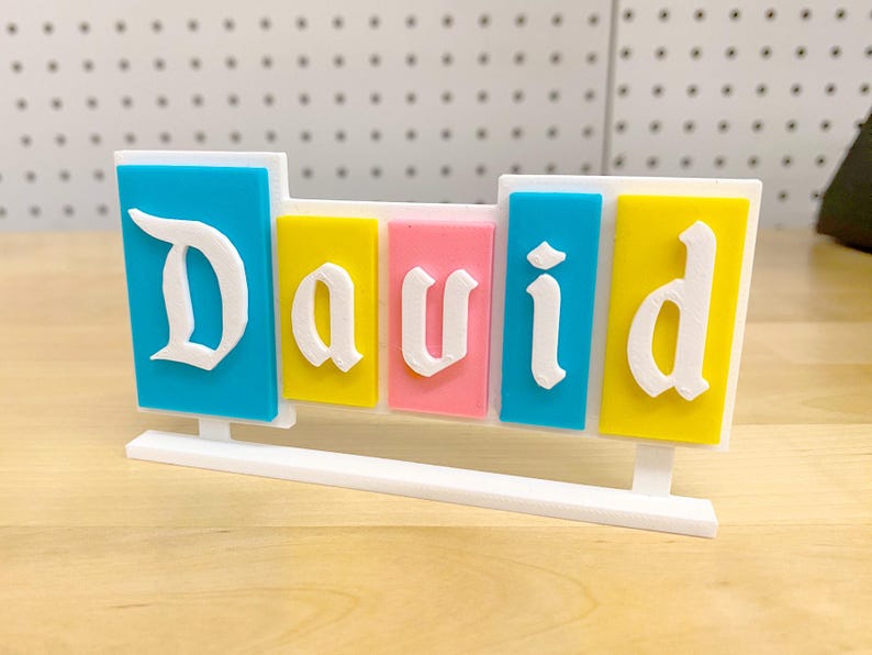 May include: A white nameplate with the name "David" spelled out in white letters on colourful blocks. The blocks are blue, yellow, pink, blue, and yellow.