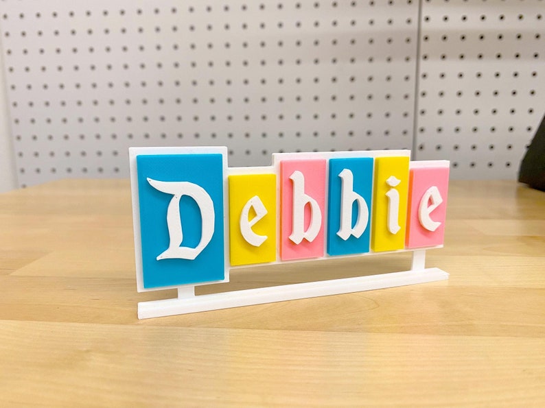 May include: A white stand with the name "Debbie" spelled out in colourful blocks. The blocks are blue, yellow, pink, and light blue.