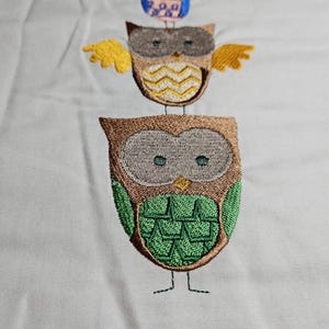 May include: Three embroidered owls stacked on top of each other. The bottom owl is green, the middle owl is yellow and brown, and the top owl is blue and pink.