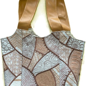 May include: A patchwork tote bag with brown leather straps and base. The bag's body features embroidered patterns in brown, tan, and light blue. The bag has a structured shape and a wide opening. The bag's measurements are in centimeters.