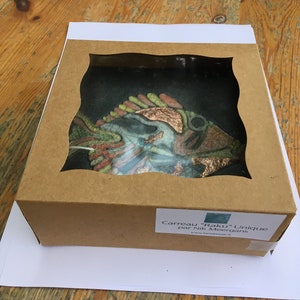 May include: A unique, handcrafted glass tile featuring a colorful fish design with copper accents. The tile is displayed in a brown cardboard box with a clear window. The text on the label reads "Carreau 'Raku' Unique par Nik Meergrans".