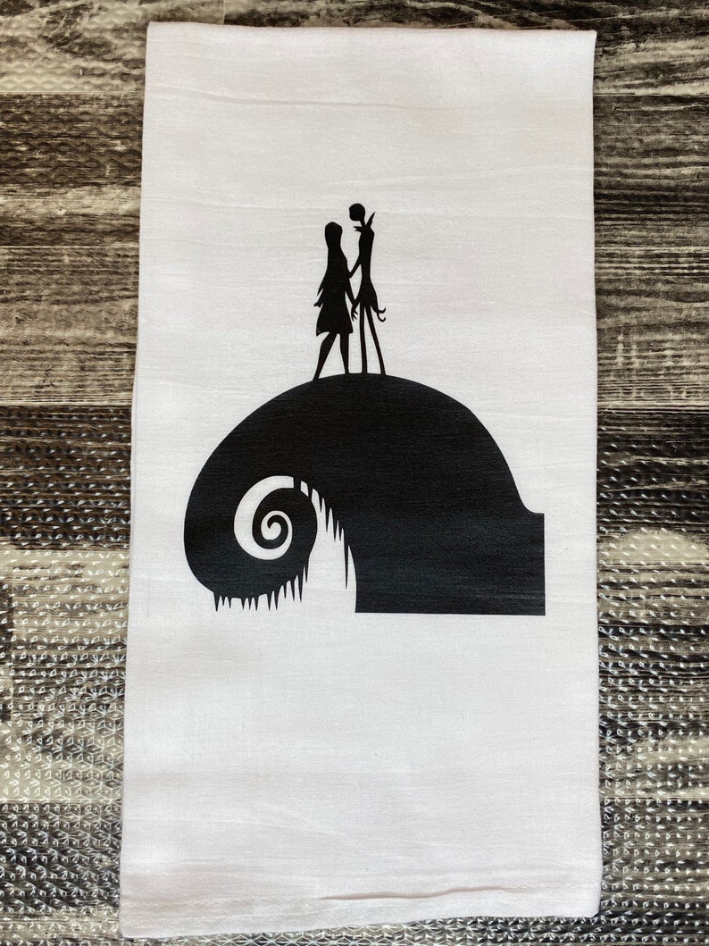 Może przedstawiać: Biały ręcznik kuchenny z czarną sylwetką Jacka Skellingtona i Sally z filmu Koszmar przed świętami stojących na czarnej sylwetce kultowego wzg&oacute;rza Halloween Town.