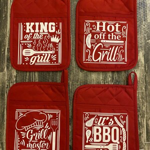 May include: Set of four red quilted pot holders with white text and graphics. The pot holders feature different grilling-themed sayings, including "King of the Grill", "Hot off the Grill", "Grill Master", and "It's BBQ Time".