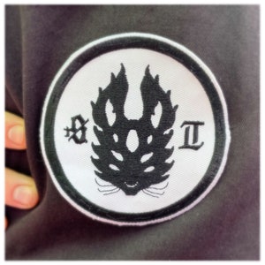 May include: A white circular patch with a black embroidered design of a creature with a flame-like head and the letters "X" and "II" on either side.