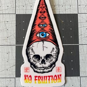 May include: A sticker featuring a skull with a red and white hat adorned with multiple eyes. The text "NO FRUITION" is displayed in yellow and orange. The sticker has a white border and is on a gray surface.